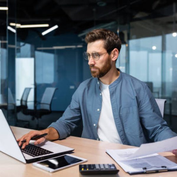 istockphoto-1473781095-612×612 A serious young man accountant, financier, analyst, auditor sits in the office at the table. He holds documents and a pen in his hands, checks accounts, finances, types on a laptop.